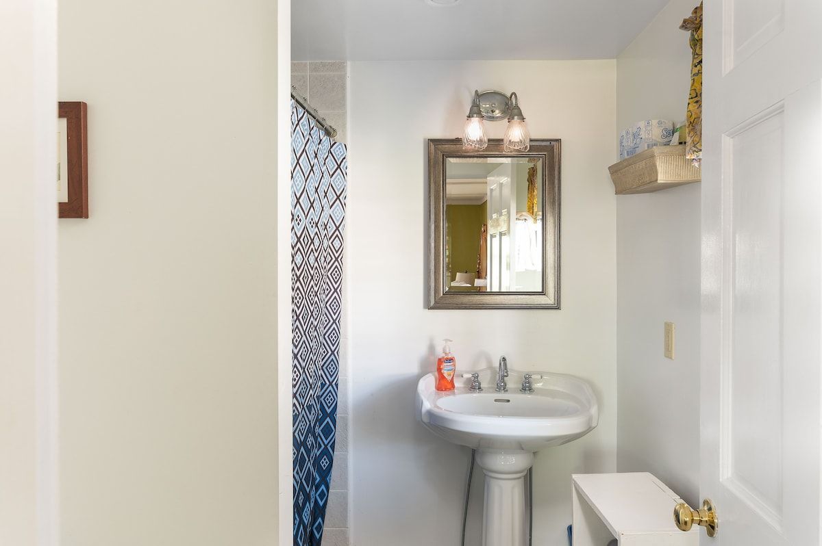 A small bathroom with a pedestal sink and mirror, featuring a soap dispenser on the counter.