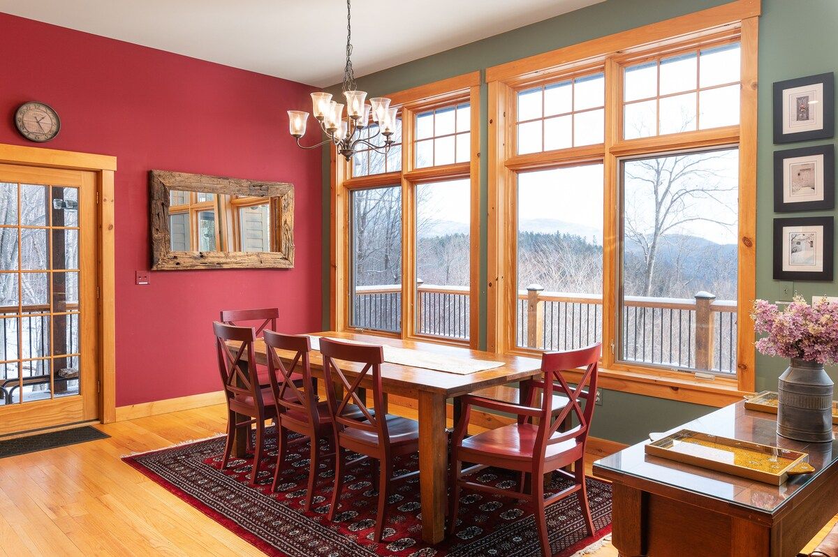 A cozy dining room with a large table and chairs, offering a scenic view of the mountains through the windows.