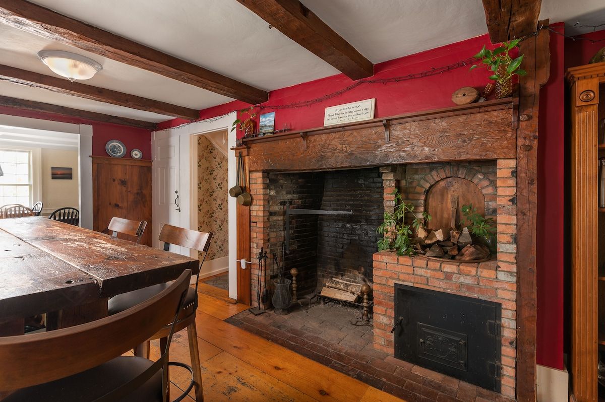 A rustic dining room with a large wooden table and chairs, featuring a brick fireplace as a focal point.