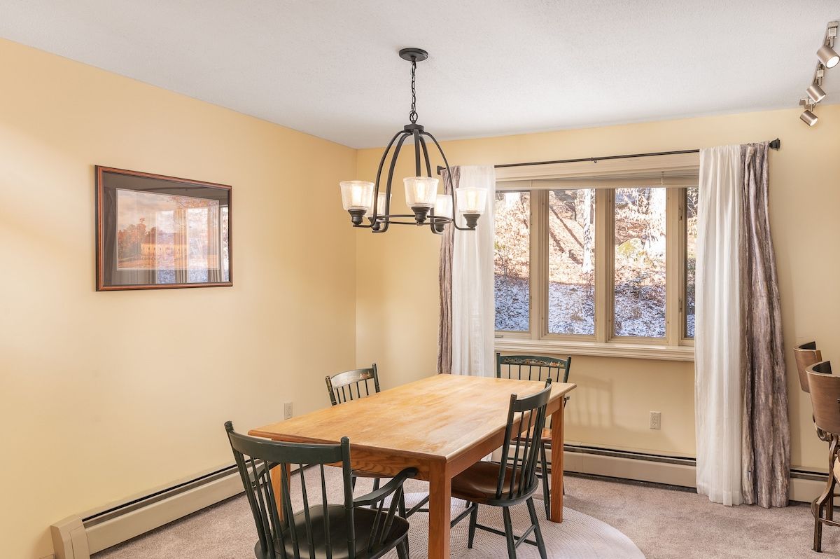 A cozy dining area with a wooden table and chairs, surrounded by cream-colored walls and a large window.