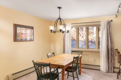A cozy dining area with a wooden table and chairs, surrounded by cream-colored walls and a large window.