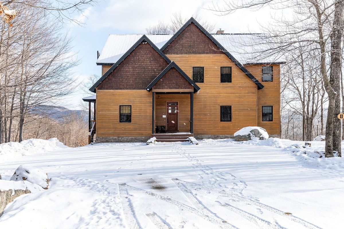 A large, two-story house with a brown exterior and snow-covered roof in a wooded area.