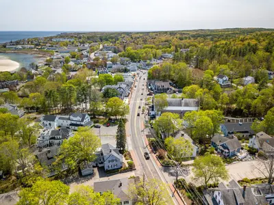 Aerial view of a small coastal town with a mix of residential and commercial buildings, surrounded by trees and a beach