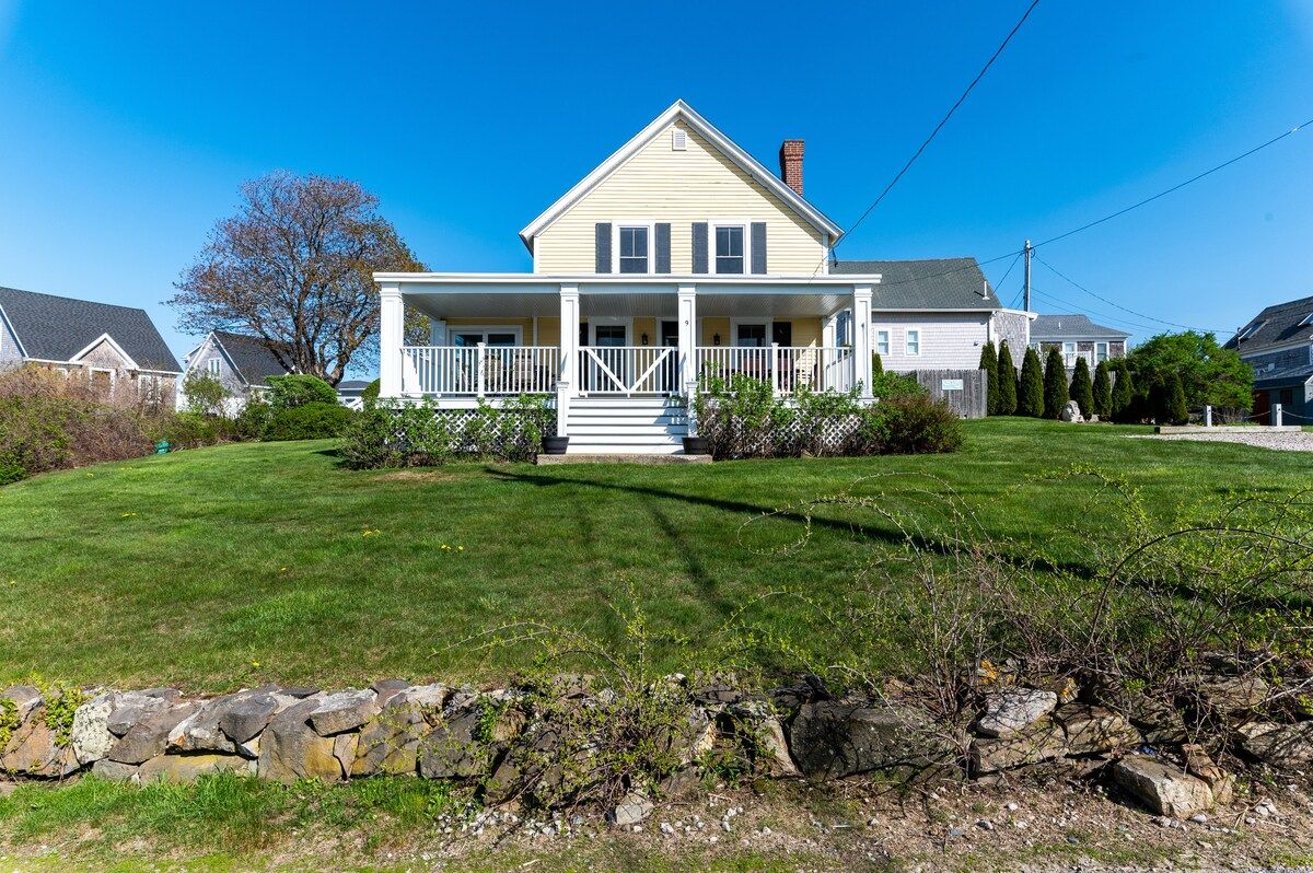 A charming two-story house with a spacious porch and a well-manicured lawn, set against a clear blue sky.
