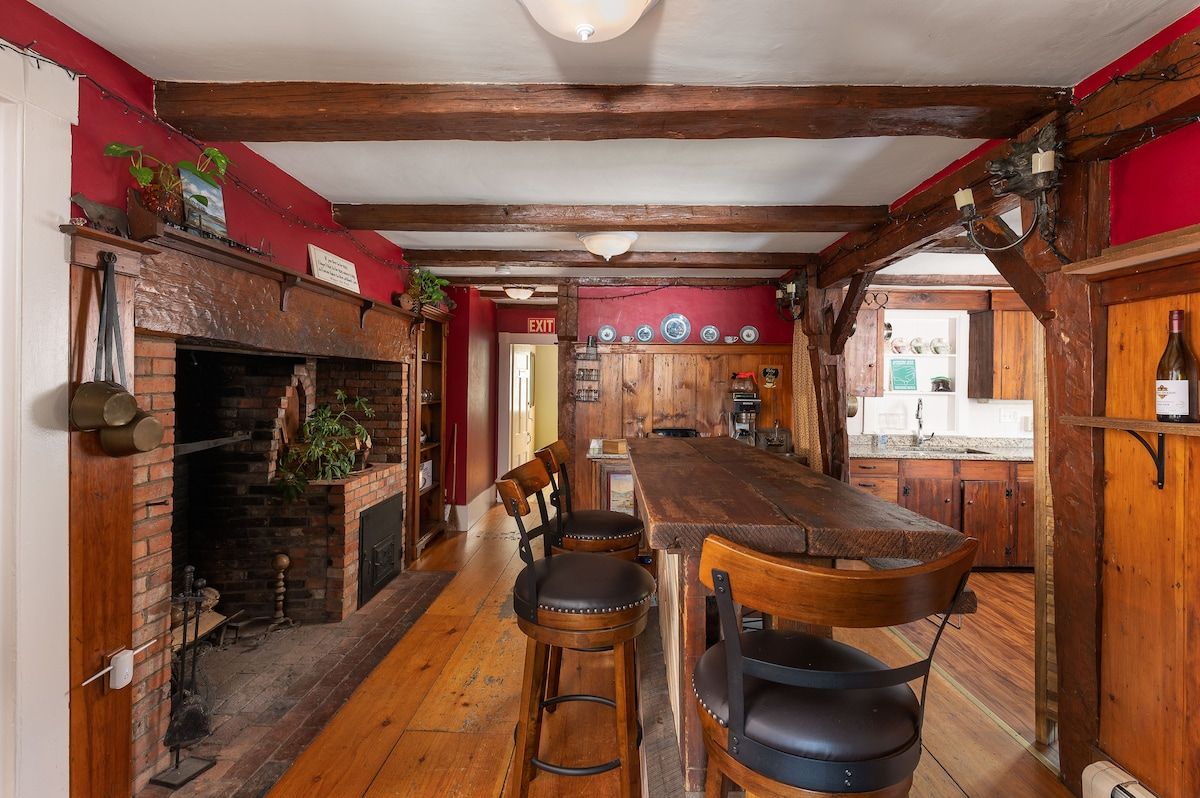 A rustic kitchen with a large wooden table and chairs, featuring a brick fireplace on the left.