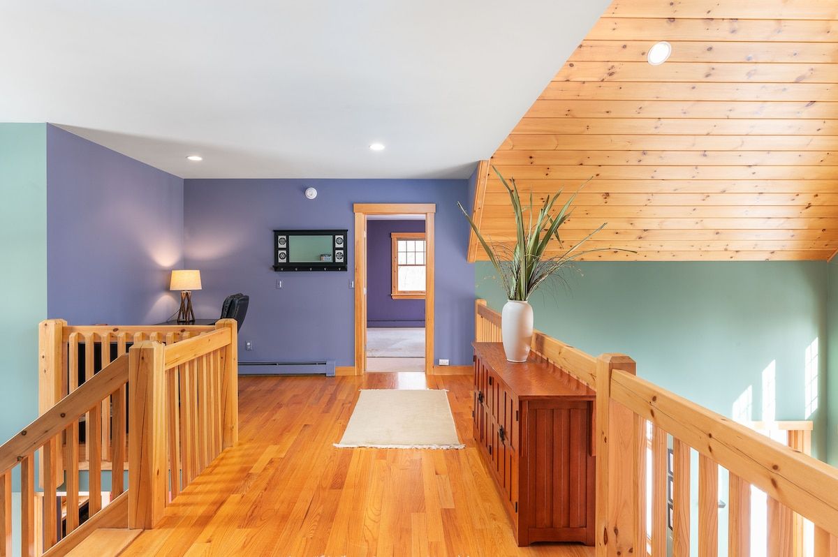 A spacious hallway with a wooden floor and railing, featuring a TV on the wall and a desk with a lamp.