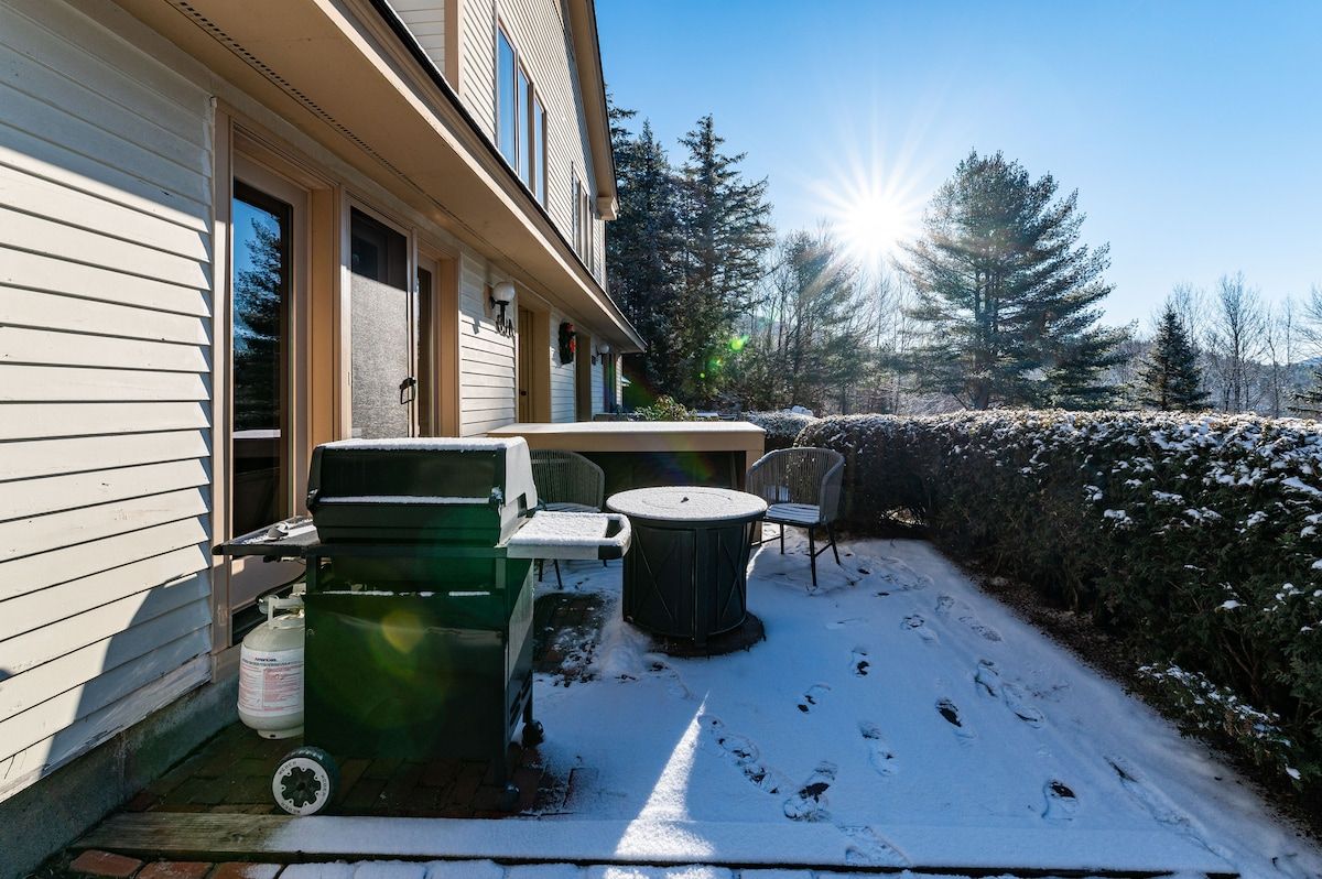 Snowy patio with a grill and table, surrounded by trees and bushes on a sunny day.