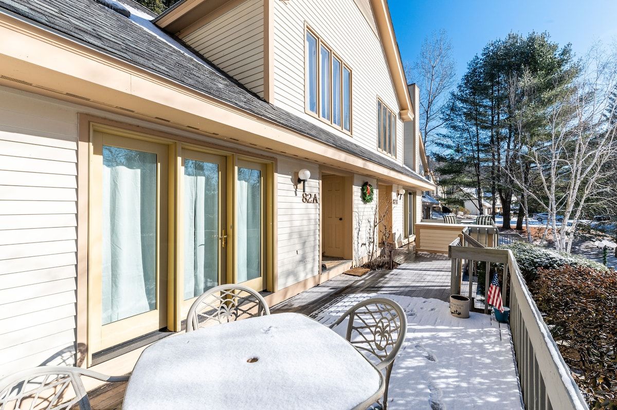 Snowy deck with a table and chairs, surrounded by trees and a blue sky.