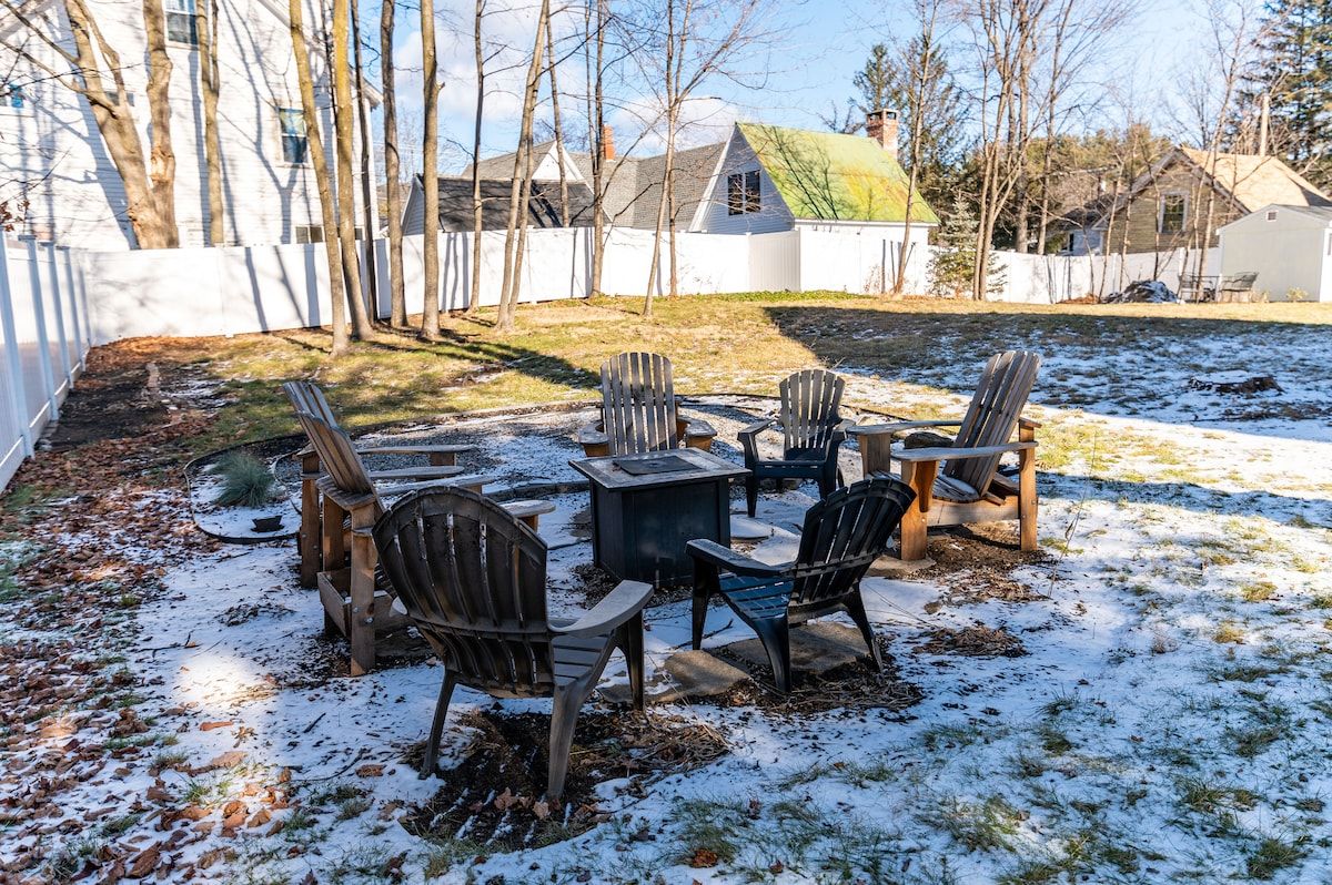 A snow-covered backyard with a fire pit and Adirondack chairs, surrounded by trees and houses in the background.