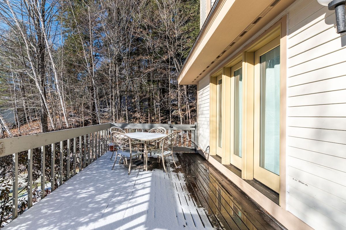 A snow-covered deck with a table and chairs, surrounded by trees and a blue sky.