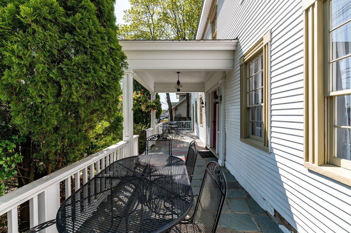 A covered porch with a black metal table and chairs, surrounded by trees and bushes.