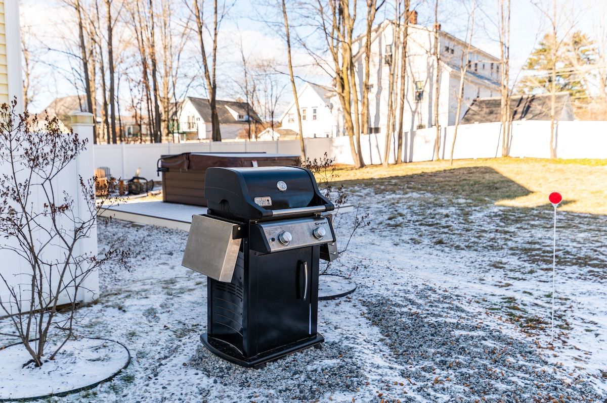 A snow-covered backyard with a grill and hot tub, surrounded by trees and houses in the background.