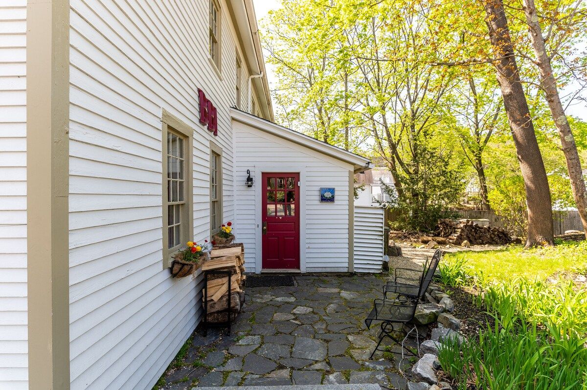 A charming white house with a red door and a cozy patio area, surrounded by trees and greenery.