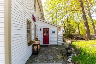 A charming white house with a red door and a cozy patio area, surrounded by trees and greenery.
