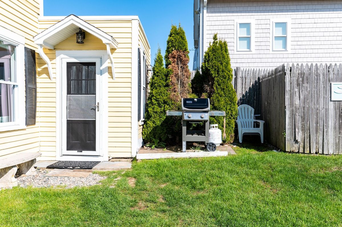 A small outdoor grill and patio area with a white Adirondack chair, surrounded by trees and a wooden fence.
