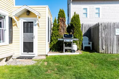 A small outdoor grill and patio area with a white Adirondack chair, surrounded by trees and a wooden fence.