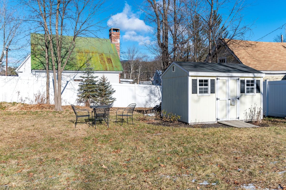 A small white shed with a gray roof and a patio set in the backyard of a house.