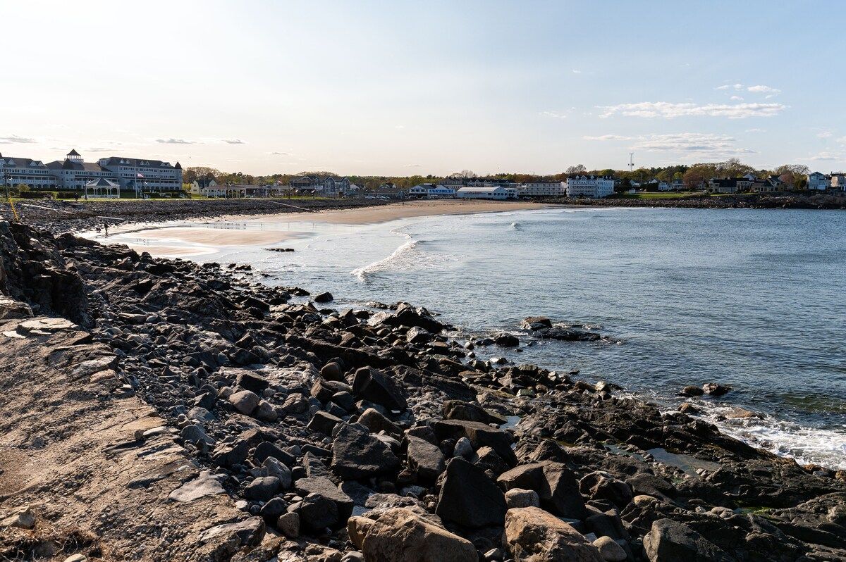 A rocky beach with a sandy shore and buildings in the background on a sunny day.