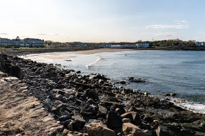 A rocky beach with a sandy shore and buildings in the background on a sunny day.