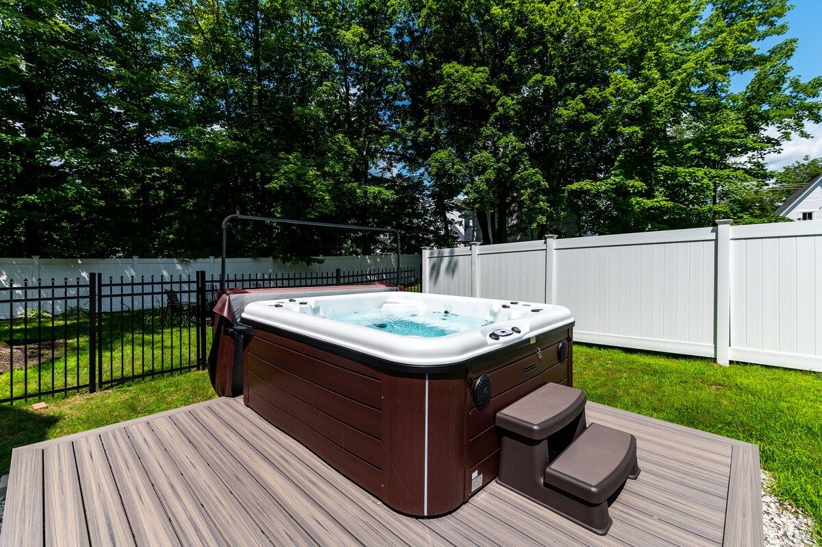 A backyard with a hot tub on a wooden deck surrounded by trees and a white fence.