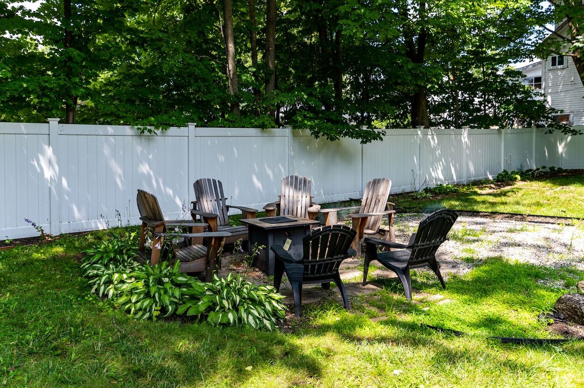 A cozy backyard seating area with a fire pit and wooden chairs, surrounded by lush greenery.