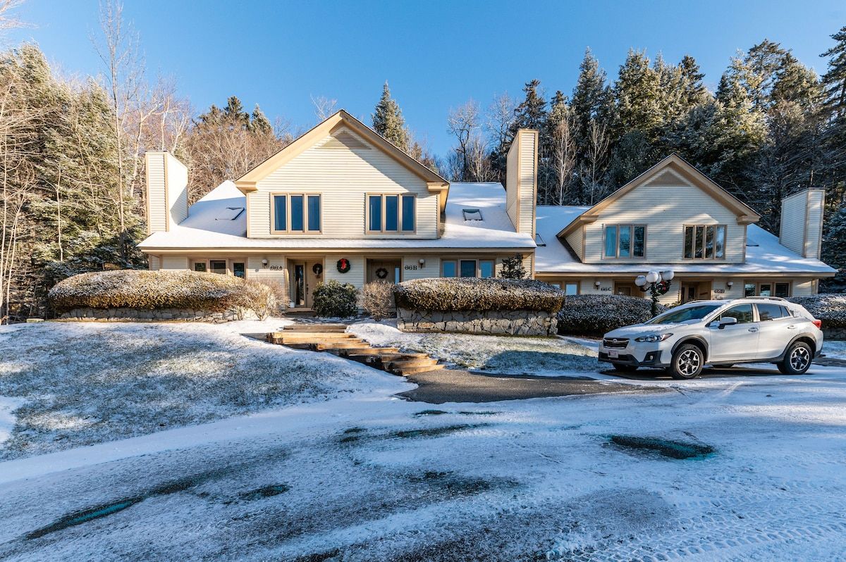 A large, two-story house with a white exterior and snow-covered roof, surrounded by trees and a clear blue sky.