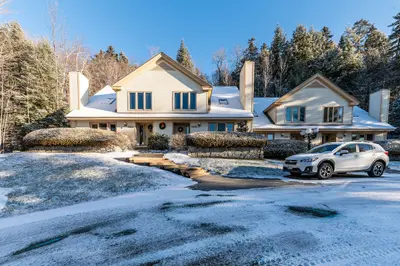 A large, two-story house with a white exterior and snow-covered roof, surrounded by trees and a clear blue sky.