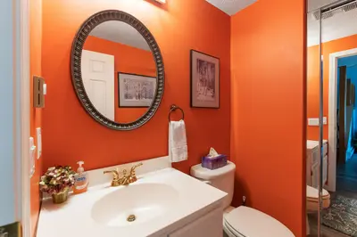 A small bathroom with a white sink and toilet, featuring an orange wall and a round mirror.