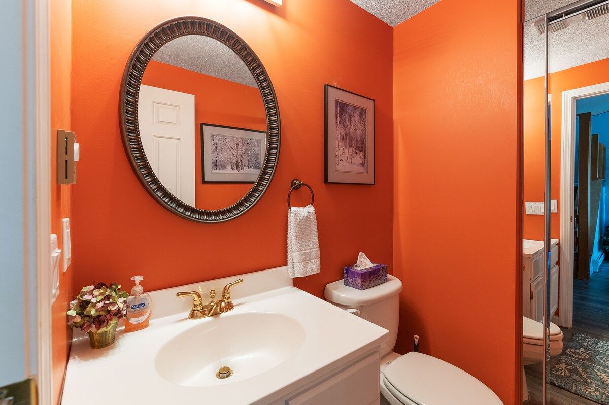 A small bathroom with a white sink and toilet, featuring an orange wall and a round mirror.